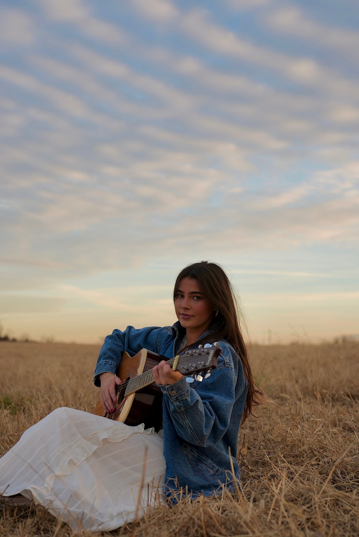 Kailey Jane sitting in a field playing guitar at sunset